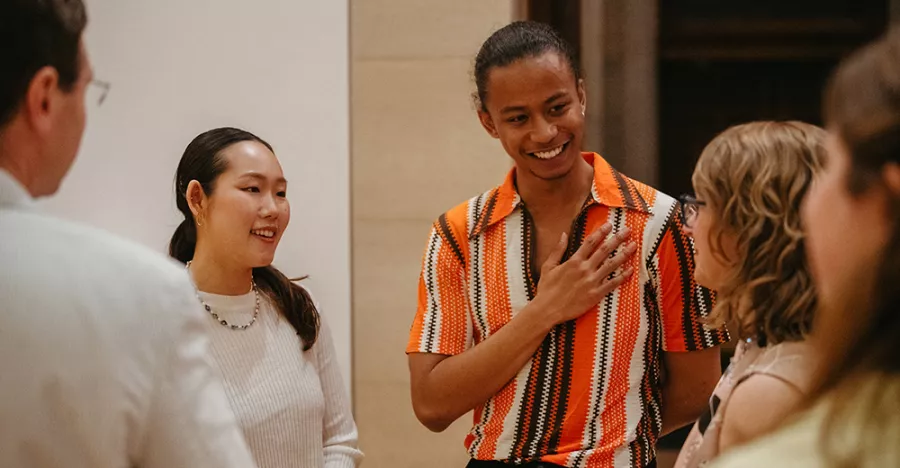Two people are standing in a circle talking to others. The person on the right is wearing orange and has their right arm over their chest. The person on the left is wearing white.