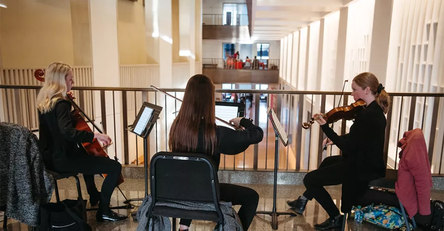 Three people hold string instruments while looking at white sheets with music. They are positioned in front of a railing.