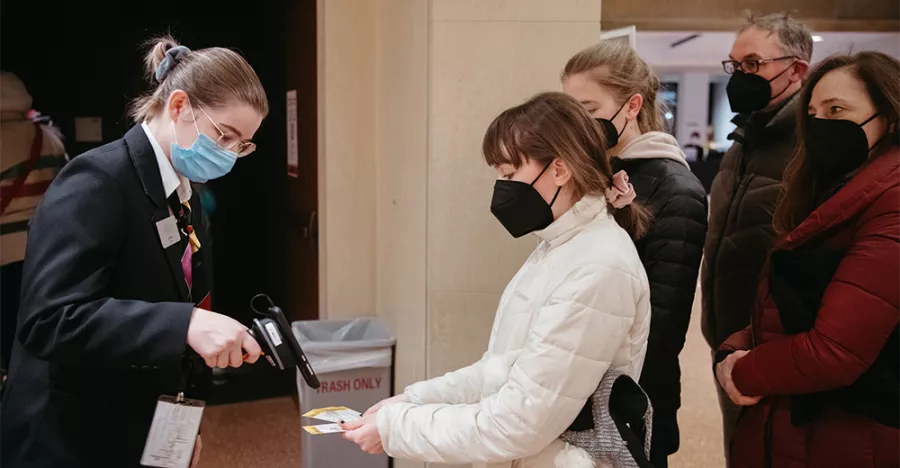 Four patrons wearing black masks face an usher who is wearing a black suit jacket and blue mask. The usher is holding a black device to scan the patrons’ tickets.