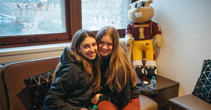 Two students wearing winter coats sitting on a couch smile for a picture. There is a UMN Mascot, Goldy Gopher, stuffed animal to their right.