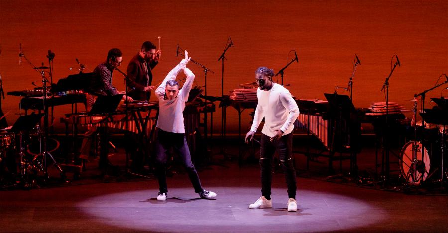 A red lit stage shows two male dancers, both wearing white longsleeve shirts and dark pants. The dancer to the left strikes a contorted pose with his arms behind his head.