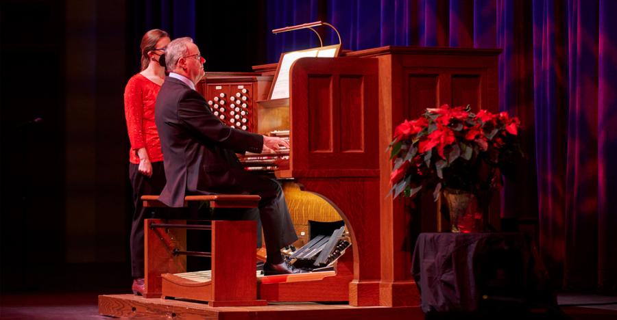 A purple and red lit stage shows a man playing a large historic organ, with a woman in a red shirt and mask standing to his left. To his right, is a large, red, poinsettia plant. 