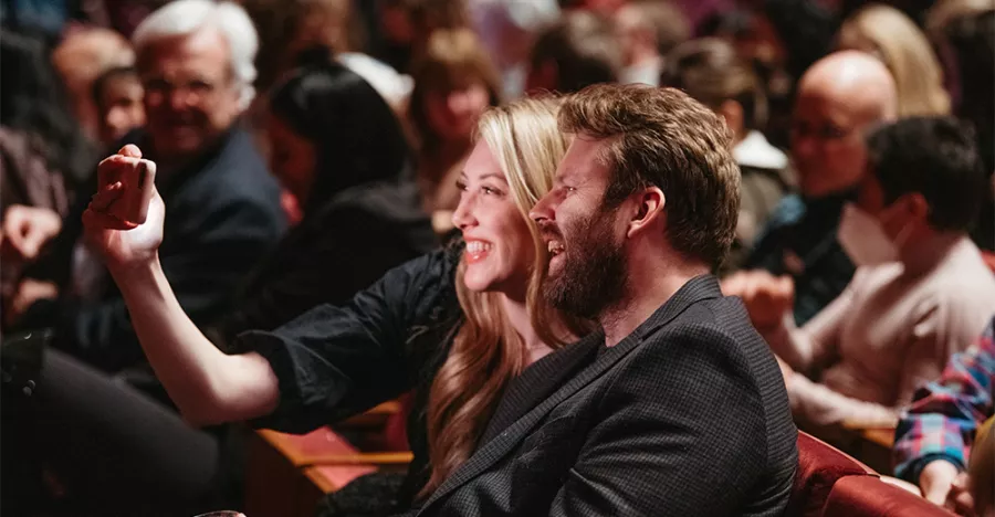 Two people smile for a selfie. The person on the right is holding their arm out with their phone to take the picture. They are sitting in maroon seats surrounded by other audience members.
