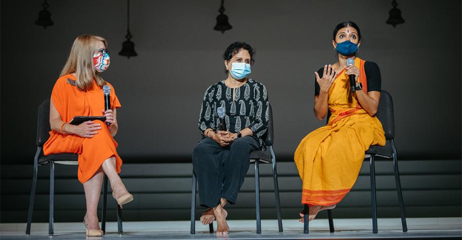 Three people sitting on chairs for a Q&A. From left to right the people are wearing an orange dress, then black pants and a black printed shirt, and an orange costume. All people are wearing masks.