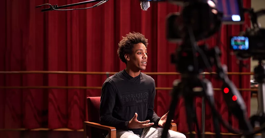 Anthony Santos sits in a chair in front of maroon curtains facing a few cameras.