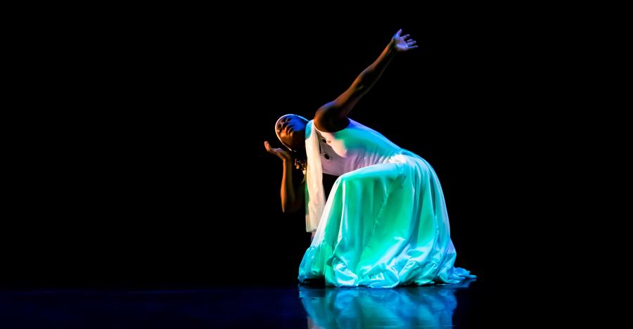 A dark stage shows a woman wearing all white, striking a pose on her knees with her arms extended. A blue and green light shines onto her white dress.