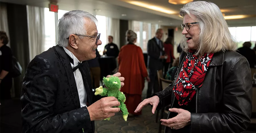 Hector Olivera and an audience member exchange conversation over a stuffed animal that is a frog. Olivera is in a black tux and the audience member is in a black leather jacket with a red scarf.