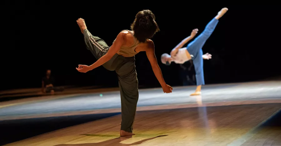 The back profile of a female dancer with short brown hair, shows her leaning backwards with her left leg extended in front of her. 