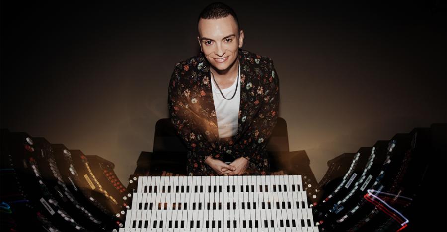 A young man wearing a patterned jacket and dark pants sits facing the camera. Before his lap are the keys white of an organ instrument. Behind the man is a dark, faded grey background.