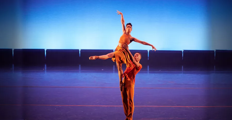 A blue lit stage shows a man holding a female dancer above his head on center stage. The female has all her arms and legs extended. 
