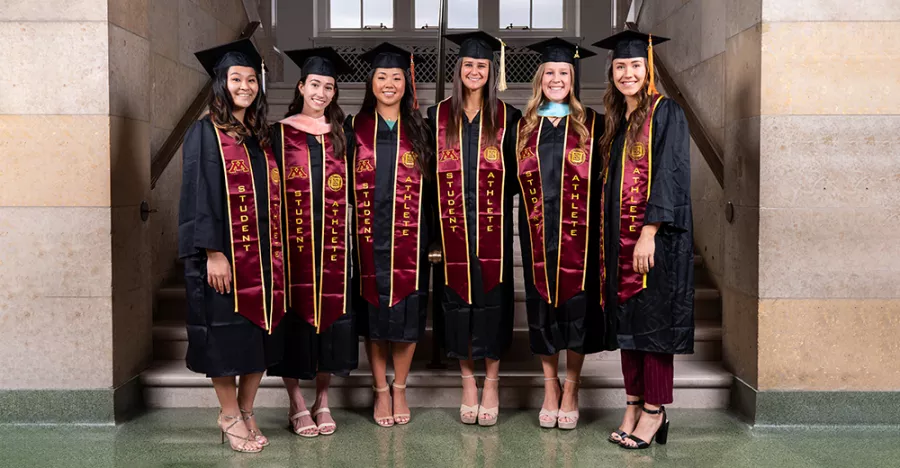 Six people stand in a marble stairway. They are wearing black robes with maroon and gold sashes.