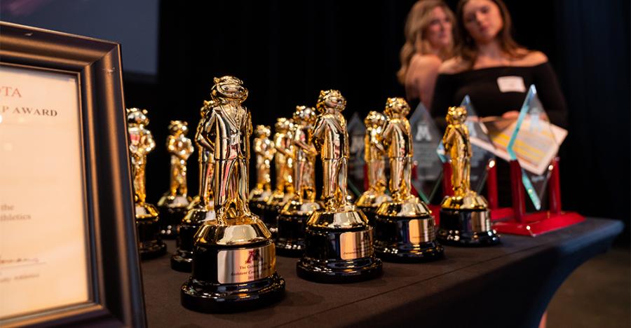 Rows of golden trophies sit on a black table. There are two people who are blurred in the background.