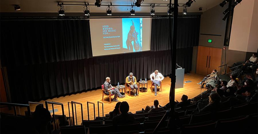 Three panelists sit on stage in front of an audience. A screen behind the panelists shows the text “ICED BODIES: ICE MUSIC FOR…” and an image of the artist on the right side.