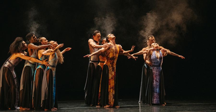 A dark stage shows three vertical lines of dancers, all striking a pose with both arms out to either side of their bodies, and their heads tilted back.