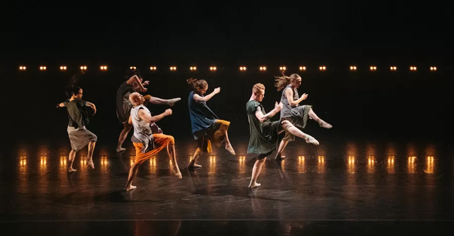 A yellow lit stage shows two horizontal lines of dancers, one in front of the other, with all dancers striking a running pose. 