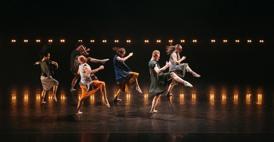 A yellow lit stage shows two horizontal lines of dancers, one in front of the other, with all dancers striking a running pose. 