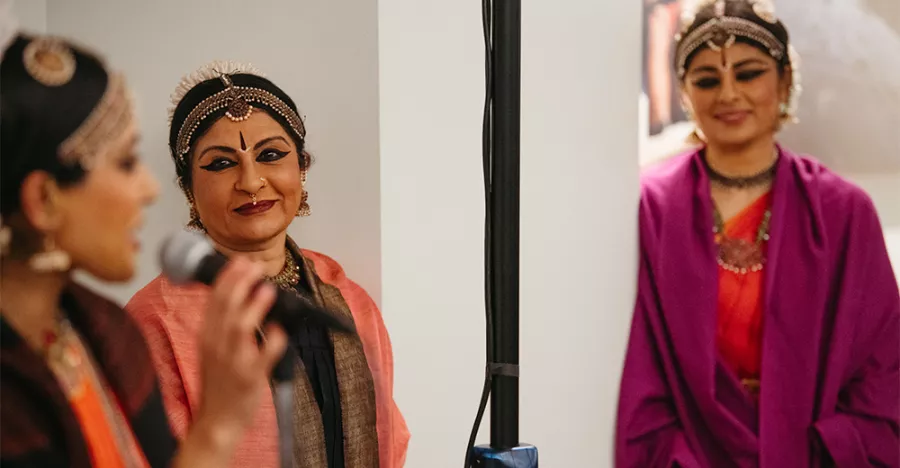 One woman speaks into a microphone while two other woman watch her with smiles on their faces.