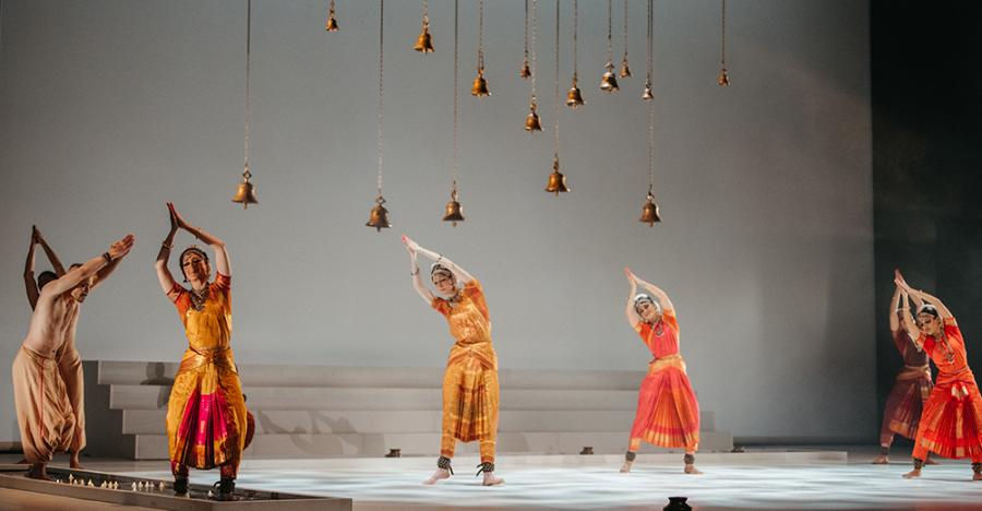 A bright white stage with a white backdrop shows a horizontal line of dancers, all wearing shades of orange, striking a post with both arms over their heads. Copper bells of varying lengths hang above their heads.