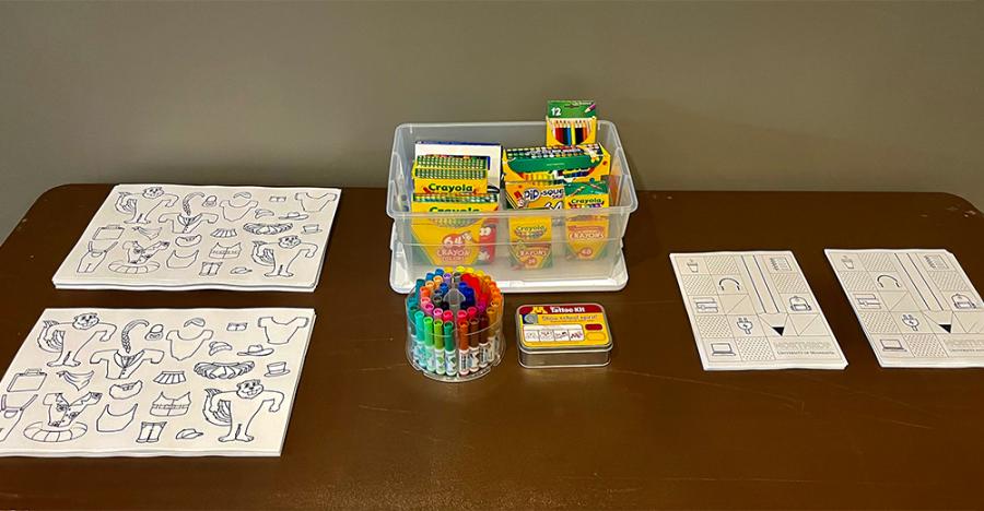 Black and white coloring sheets with gophers and various clothing items sit on a brown table along with crayons and markers. There is another coloring sheet with icons like pencils, headphones, and computers.