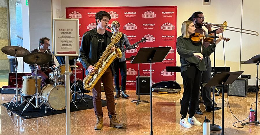 Five people stand on a marble floor with a red banner in the background playing instruments such as the drums, saxophone, guitar, trombone, and violin.