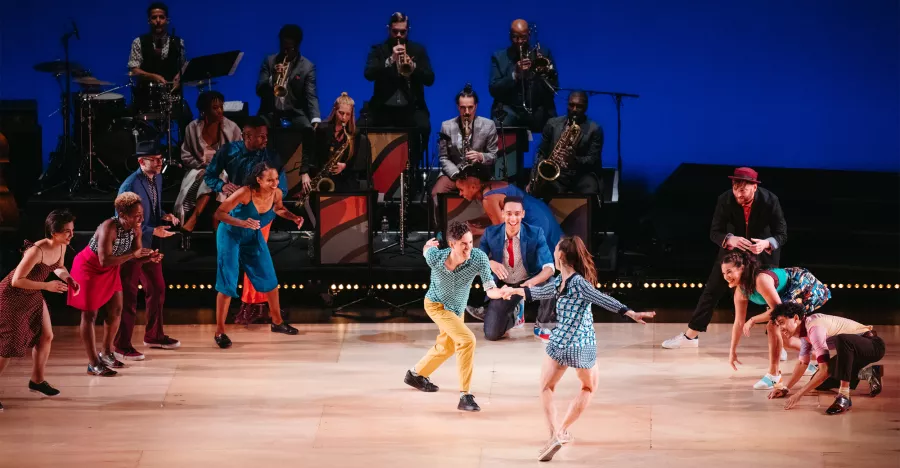 A large group of dancers wearing bright colors, form a circle that outlines a man and a woman dancing hand in hand on center stage. The outlined dancers are all clapping ang watching the man and woman.