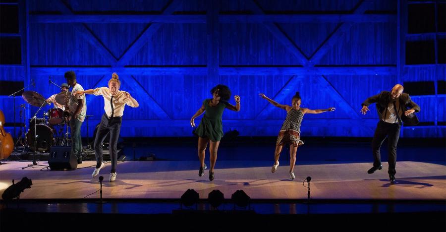 A blue lit stage shows a horizontal line of dancers all tap dancing, with both arms out to either side of their bodies.