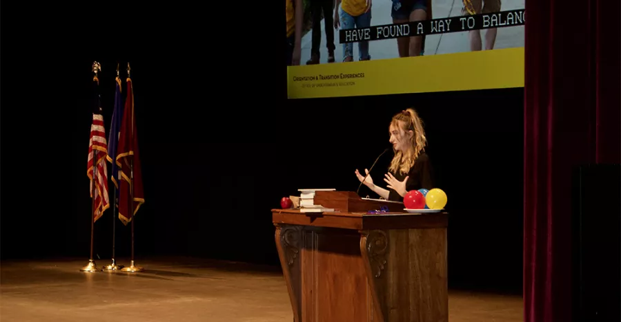 A female speaker stands at a wooden podium delivering a speech. There are three flags on the left side of the image and a projector screen in the back that shows the legs of three University students and a caption that reads, “Have found a way to balance.” 