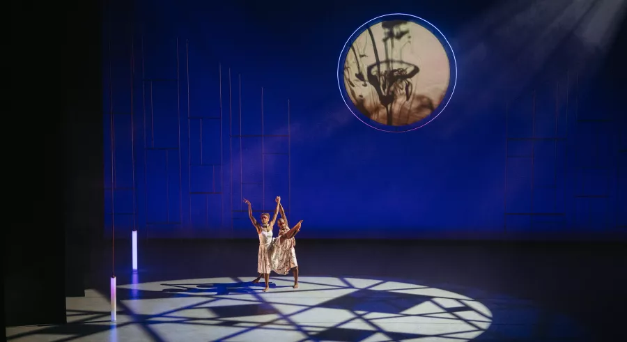 A dancer in a white, patterned dress lifts their leg as a second dancer also dressed in a similar outfit holds their lifted leg and extended hand. A circular image with cream and black colors is seen on the wall behind the dancers. 