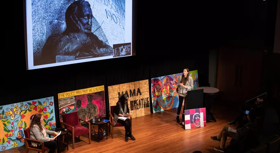 One person stands behind a podium, discussing a photo of a monument on a large screen. The other two people on stage sit in chairs in front of various murals.
