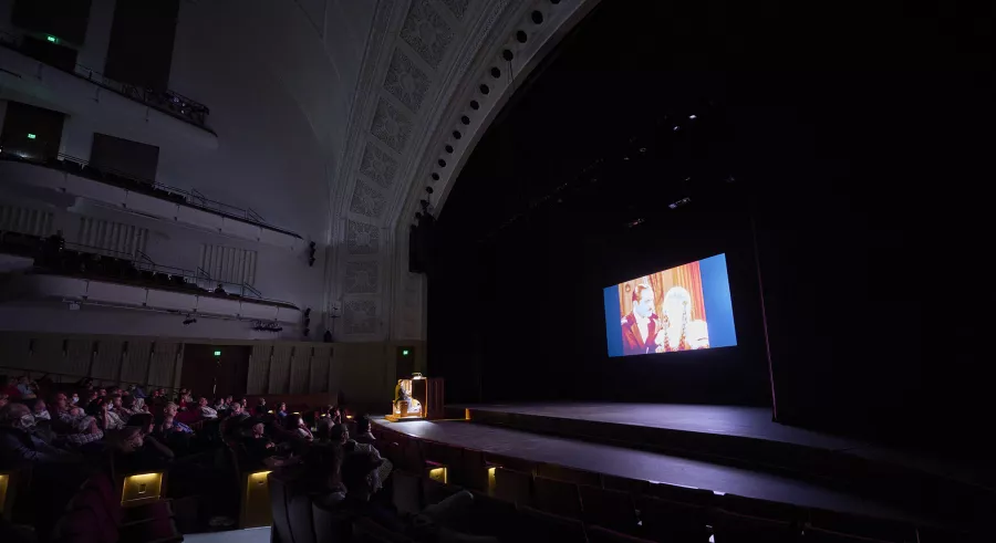 An audience watches a film onstage in a darkly-lit auditorium while an organist plays in the center.