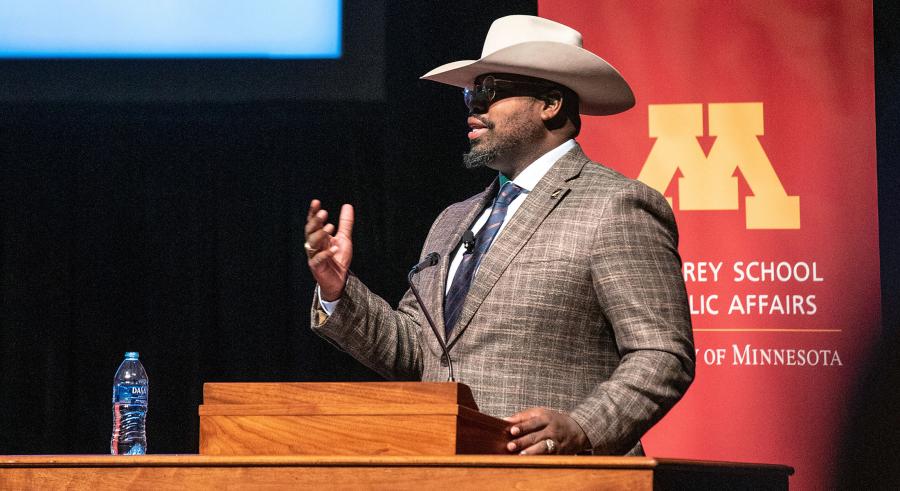 Marshall Johnson stands onstage behind a podium in a suit and cowboy hat, talking to the audience.
