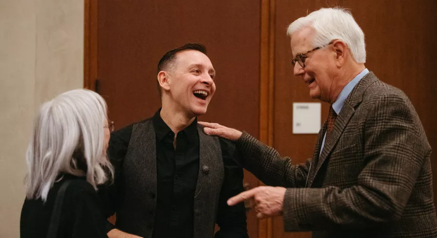 Three people laugh and talk in a small group.One has one hand on the shoulder of another and points with the other hand.