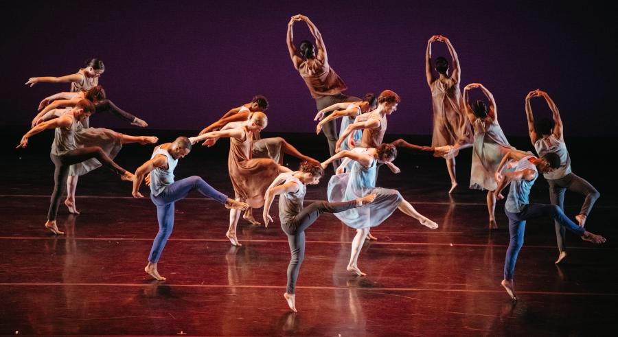 A group of dancers dressed in a variety of muted colors perform onstage. The bodies of the dancers towards the front are turned to the side as they extend one of their legs forward with their arms behind themselves. Dancers behind them face the back of the stage with hands interlocked above their heads and bent, lifted knees.