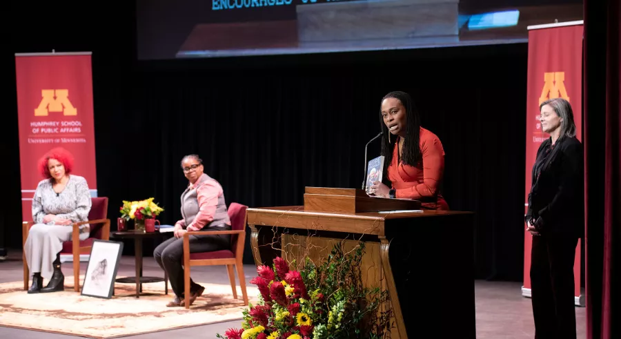 Two people sitting onstage while one stands behind the podium holding a book, with one more person standing behind the speaker. 