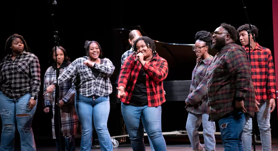 A choir of eight people performing onstage wearing an assortment of plaid shirts, smiling. One singer in front is holding a microphone and singing. 