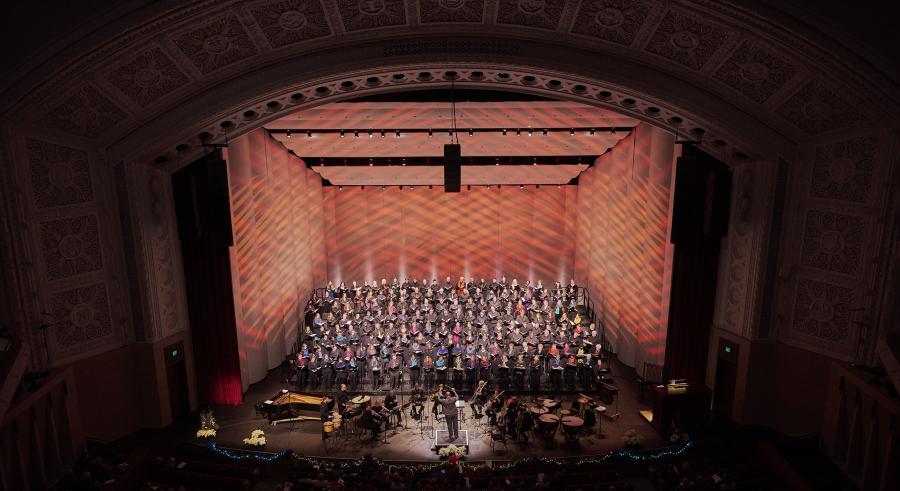 A large choir performs on risers in an auditorium filled with red and orange. A conductor stands in front of the choir, leading them and instrumentalists while an audience watches.