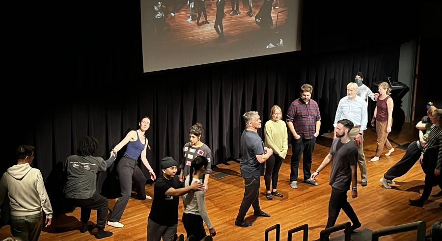 Sixteen people stand scattered around the stage, participating in various movement exercises with one another.