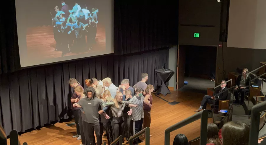 A group of people stand in a circle facing outwards, linking arms at the elbow. Audience members are seated and watching, and a projected screen hangs behind the stage, displaying a live feed of the presentation. 