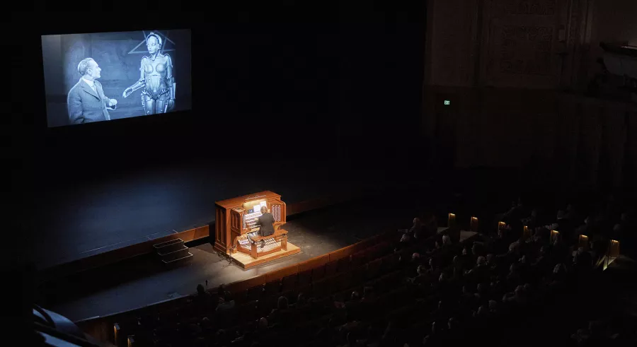 An audience watches a film onstage in a darkly-lit auditorium while an organist plays in the center. 