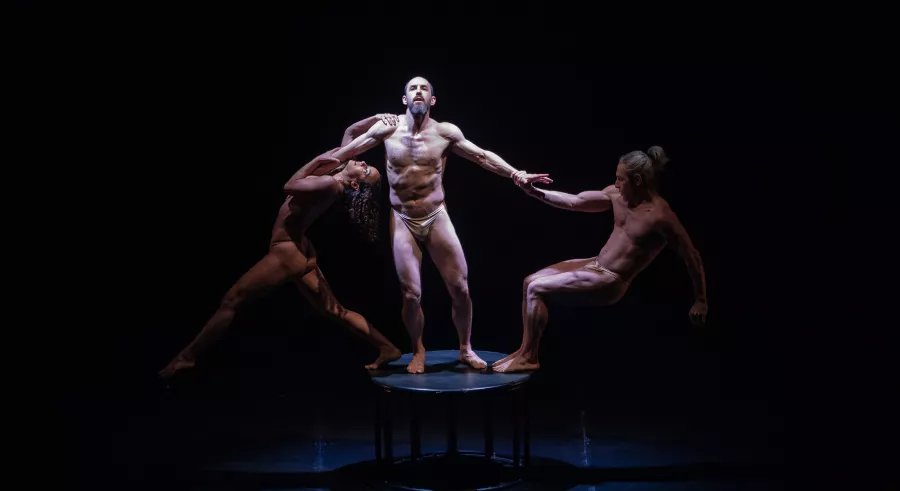 Three dancers link hand, one stands tall in the center, with a white spotlight shining on them, the two on the sides crouch low and are in half-shadow.