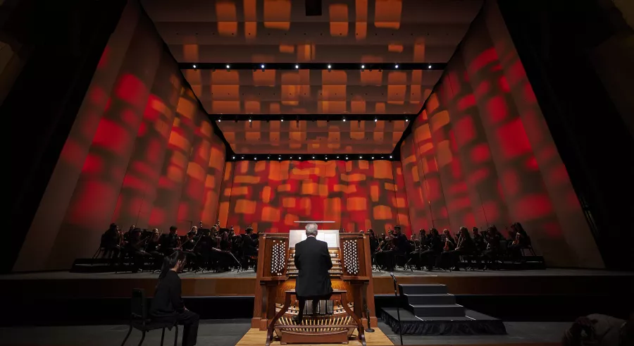 Dean Billmeyer plays the organ in front of an orchestra also dressed in black. Behind the orchestra onstage, the walls are lit with red, rectangular-shaped light.