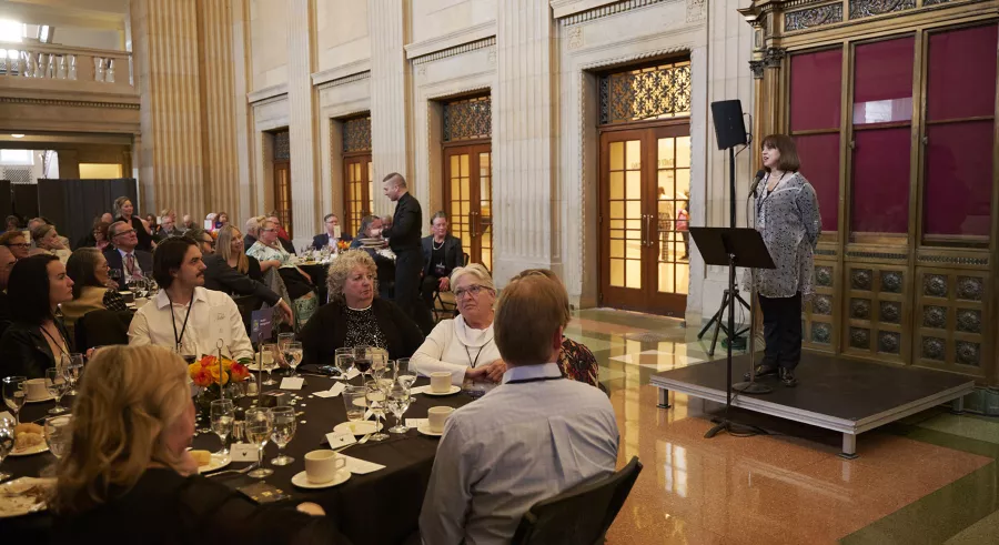 A speaker holding her hands behind her back stands on a raised platform and addresses a crowd of people sitting at tables.