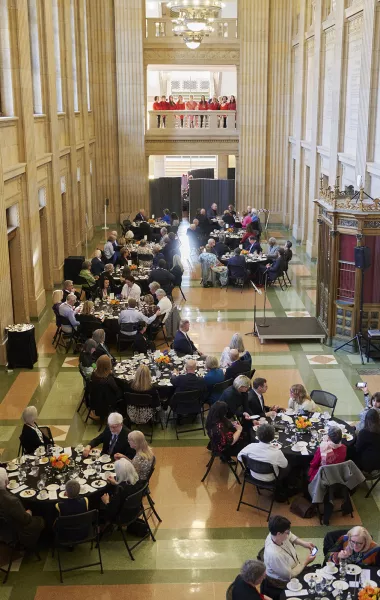 People in formal attire sit and stand around tables while talking to one another in a large hall.