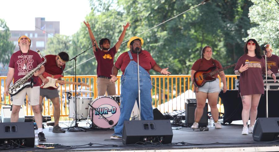 Seven musicians on stage performing on the Northrop Plaza. They are each wearing maroon and playing various instruments. Nur-D stands in the middle in front of a microphone.