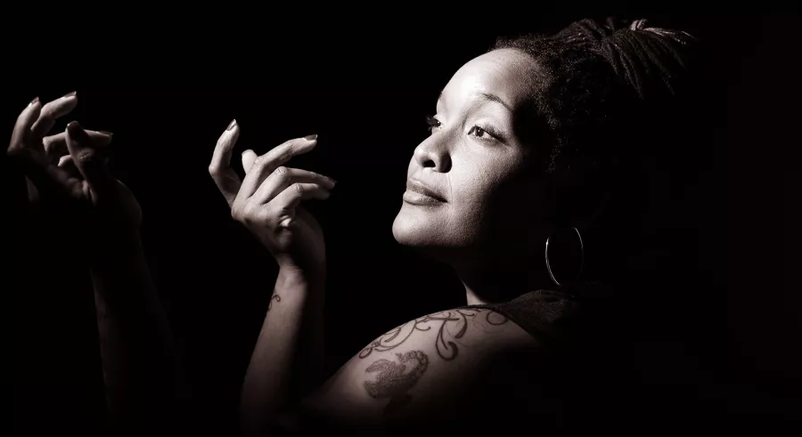 Vocalist PaviElle in front of a black background. Her side profile is illuminated by a white light, and her hands are raised in front of her. 