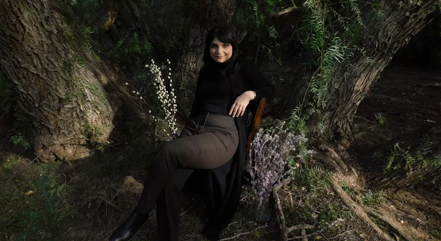 Sarah sits outside in a chair by a tree smiling at the camera