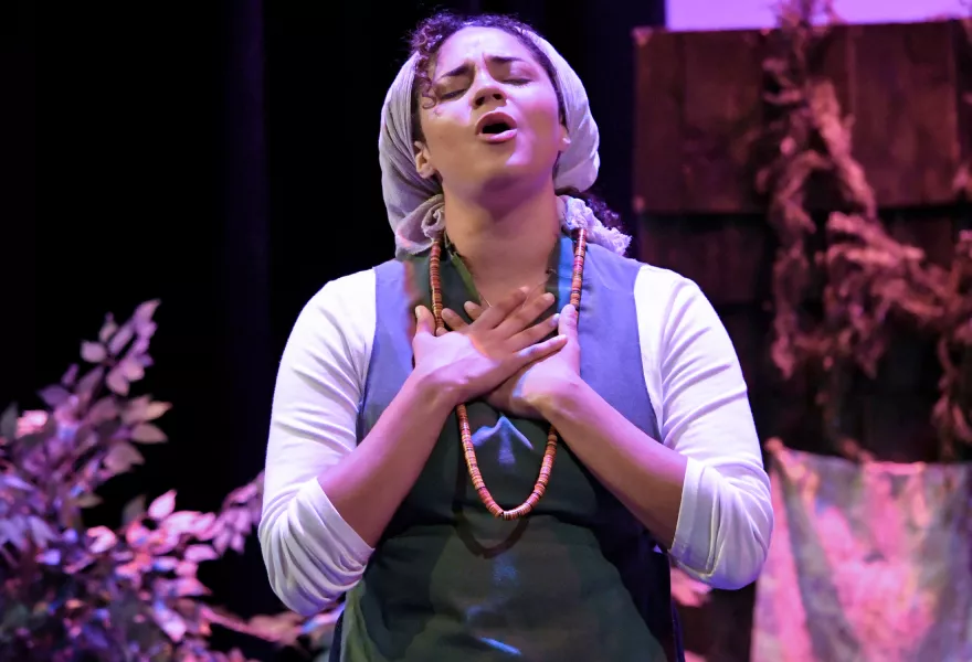 Woman in purple dress and headscarf sings, head raised, eyes closed, hands over her heart.