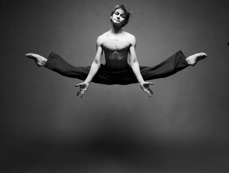 Daniil wears black pants in center-split jump high above the floor, arms down and open.