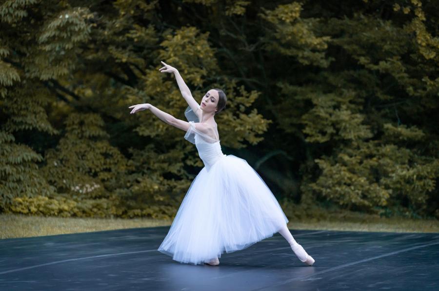 A dancer in a white dress gracefully raises her arms and points her foot. 