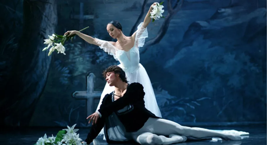 A man sits on the stage with his legs stretched out. A woman in a white dress stands behind him with her arms spread wide.